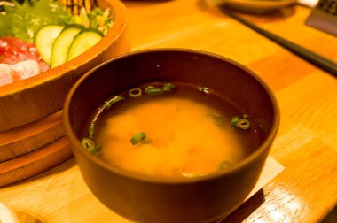 Dilimlenmiş yeşil soğanla süslenmiş dumanı tüten miso çorbası ahşap bir masada geleneksel ahşap bir sashimi küvetinin yanında duruyor. Sıcak aydınlatma rahatlatıcı, kırsal atmosferi vurgular.