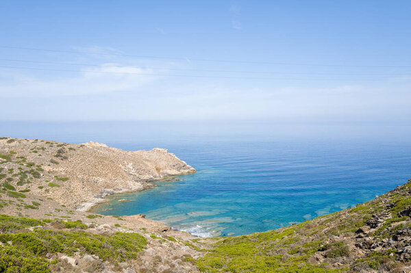 Bright midday sun illuminates a rugged coastline with rocky slopes and vibrant green shrubs leading down to a crystal-clear turquoise bay in Asinara National Park, Sardinia. The calm Mediterranean Sea stretches to the horizon under a cloudless blue