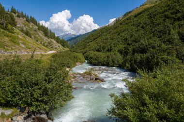 Açık alp nehri yoğun yeşil çalılar ve kayalık yamaçlar arasında enerjik bir şekilde akar, dolambaçlı bir yol ve dalgalı mavi bulutların altında uzak tepeler.