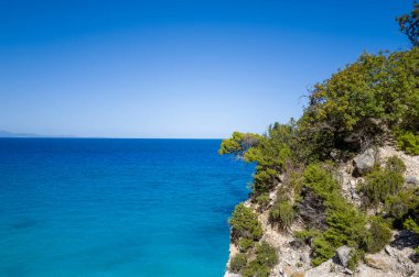 Güneşli Akdeniz bitki örtüsü, Sardunya 'nın Baunei kenti yakınlarındaki turkuaz ve derin mavi denizin üzerindeki kayalık bir uçuruma tutunur. Açık gökyüzü ve canlı kıyı renkleri ferahlatıcı, sakin bir yaz atmosferi yaratır..