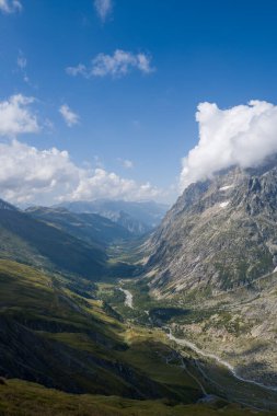Val Ferret 'in dolambaçlı bir nehri vadi tabanını, dik kayalık dağları ve mavi gökyüzünde sürüklenen dramatik bulutları süpüren görüntüsü..