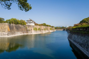 Geleneksel bir Japon şatosu devasa taş duvarların üzerinde duruyor, Osaka 'daki berrak mavi gökyüzünün altında geniş, sakin bir hendeğe bakıyor. Bereketli yeşillik ve sakin su huzurlu, güneşli bir manzara yaratır..