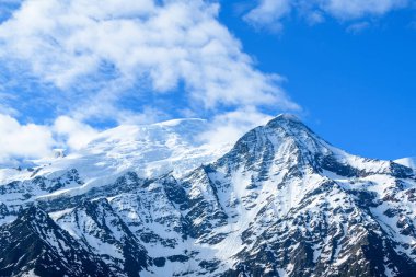 Parlak güneş ışığı, Mont Blanc ve Aiguille du Gouter 'ın engebeli, karlı yamaçlarını, dağınık bulutlarla dolu canlı mavi bir gökyüzünün altında vurgular. Dramatik alp manzarası ihtişam ve el değmemiş bir yaban hayatı barındırıyor..