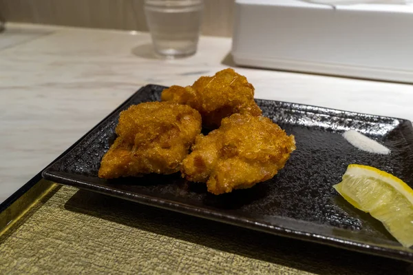 Siyah seramik tabakta servis edilen üç parça altın kızarmış tavuk, limon dilimi ve küçük bir porsiyon tuz, minimalist bir restoranda dokulu bir tepside..