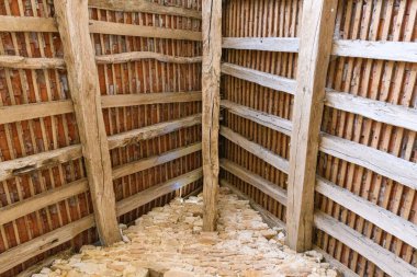 Korunmasız ahşap kirişler tarihi bir binanın iç çatı yapısındaki kil kiremitleri destekler. Yıpranmış ahşap ve pürüzlü duvarcılık geleneksel inşaat tekniklerini vurgulayan dokulu, geometrik bir kalıp oluşturuyor..