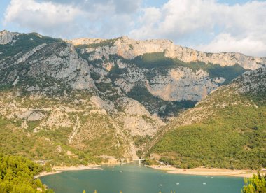 Canlı turkuaz su dramatik kireçtaşı tepelerinin altında kumlu kıyılarla buluşur ve Verdon Boğazı 'nda yemyeşil tepeler oluşur. Güneş ışığı manzaranın üzerine yumuşak gölgeler bırakır..