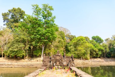 Merkezi taş heykel, yemyeşil ağaçlar tarafından çerçevelenmiş ve sakin gölete yansıtılan Kamboçya Angkor bölgesindeki Neak Pean Tapınağı 'nda suyun üzerinde yükselir. Ilık gün ışığı, yıpranmış oymaları ve huzur ortamını vurgular.