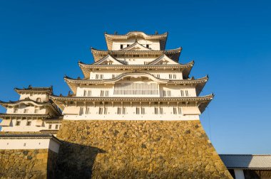 Himeji Kalesi 'nin merkezinin doğrudan manzarası katmanlı beyaz duvarlar, süslü çatılar ve parlak mavi gökyüzünün altında belirlenmiş altın bir temel..