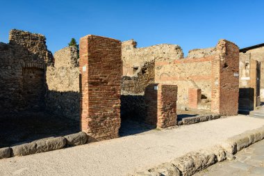 Antik tuğla sütunlar ve yıpranmış taş duvarlar İtalya 'nın Pompeii kentindeki arkeoloji sahasında asfalt bir cadde boyunca duruyor. Parlak güneş ışığı, açık mavi gökyüzünün altındaki pürüzsüz dokuları ve muhafaza edilmiş duvarları vurgular..