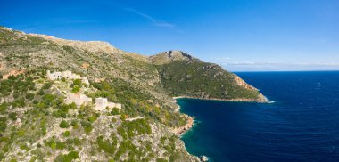 Yunanistan 'ın Mani yarımadasındaki canlı bir mavi körfezin üzerinde, engebeli dağlar ve yemyeşil Akdeniz bitkileriyle çevrili güneşli bir tepede tünemiş uzak bir taş köyünün panoramik hava manzarası.