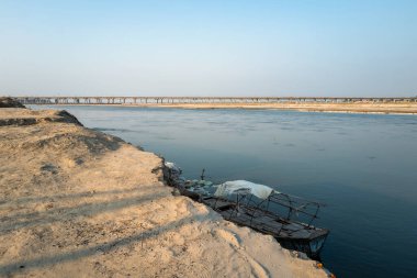 Payagraj, Hindistan 'da sakin sulara bakan kumlu bir nehir kıyısında yer alıyor. Ufuk boyunca uzanan uzak köprü berrak gün ışığında, sessiz ve geniş bir manzara yaratır..