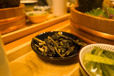 Turşulu yapraklı yeşilliklerle dolu siyah bir seramik kase sıcak ortam ışıkları altında geleneksel Japon yemekleriyle çevrili ahşap bir tepside oturur. Görüntü, sebzelerin parlak dokusunu ve toprak tonlarını vurguluyor..