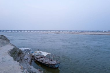 Yıpranmış ahşap bir tekne, duayraj 'daki Ganj Nehri' nin kumlu kıyısı boyunca uzanır ve sisli bir gökyüzünün altında sakin bir su üzerinde uzanan uzun beton bir köprü vardır..