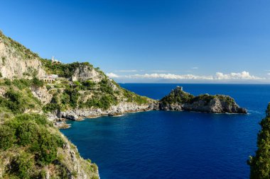 İtalya 'nın Capo di Conca kentindeki derin mavi suların üzerinde yükselen gür Akdeniz bitkileriyle kaplı kayalıklar. Tarihi bir sahil gözetleme kulesi, canlı gökyüzü ve Amalfi kıyısı boyunca ışıl ışıl deniz tarafından çerçevelenmiş küçük bir promoner üzerinde duruyor..