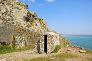 Cap de Carteret 'teki turkuaz denizin üzerindeki aşınmış kayaya karşı küçük taş yapı duruyor. Parlak güneş ışığı sert dokuları ve açık, sakin kıyı manzarasını vurgular.