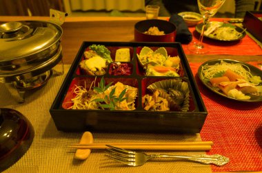 Japon aperatifleri, sebzeleri ve garnitürleriyle dolu laktüeli bir bento kutusu sıcak ortam aydınlatmasında sıcak bir tencere, sıcak bir tencere ve cam süs eşyasının yanında dokuma bir mindere yerleştirilmiştir..