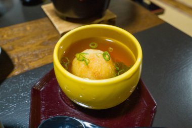 Bol yağda kızartılmış bir tofu topu, dilimlenmiş yeşil soğan ile süslenmiş ve parlak sarı bir fincanda servis edilen şeffaf bir dashi çorbasının içinde oturur. Tabak, yumuşak bir tepside sunuluyor. Sıcak tonlar ve ince Japon yemeklerinde davetkar dokular vurgulanıyor.