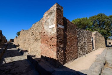 Antik tuğla ve taş duvarlar İtalya 'nın Pompeii kentindeki arkeolojik bölgede keskin bir sokak köşesi oluşturuyor. Sahne desenli duvarcılık, derin gölgeler ve bu Roma şehrinin tarihi şehir planının altını çizen canlı bir mavi gökyüzü içeriyor..
