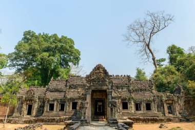 Preah Khan tapınağının yıpranmış taş cephesi Kamboçya Angkor bölgesinde soluk mavi bir gökyüzünün altında duruyor. Karmaşık oymalar, açık pencereler ve koruyucu heykeller yemyeşil ağaçlar ve kuru zemin tarafından çerçevelenerek tarihi eserlerin altını çiziliyor.
