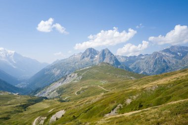 Engebeli dağ sırtları ve yuvarlanan yeşil yamaçlar parlak mavi gökyüzünün altında dolambaçlı yollar ve Mont Blanc yakınlarındaki Chamonix vadisinde dağınık bulutlar..