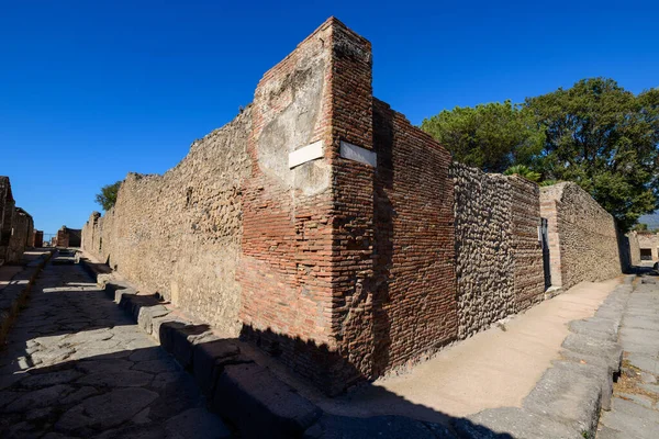 Antik tuğla ve taş duvarlar İtalya 'nın Pompeii kentindeki arkeolojik bölgede keskin bir sokak köşesi oluşturuyor. Sahne desenli duvarcılık, derin gölgeler ve bu Roma şehrinin tarihi şehir planının altını çizen canlı bir mavi gökyüzü içeriyor..