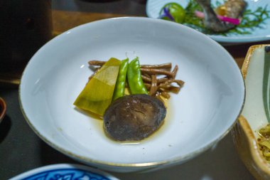 Beyaz bir seramik kasede kaynamış bir shiitake mantarı, yeşil fasulye, bambu yaprağı ve hafif bir et suyunda yabani dağ sebzeleri bulunur. Yemek geleneksel bir Japon yemek masasında toprak renkler ve ince dokularla sunuluyor..