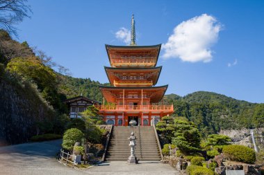 Japonya 'nın Wakayama kentindeki Kumano Nachi Taisha' daki canlı turuncu bir pagoda 'nın önünde heykel yeşilliği boyunca uzanan taş adımlarla. Tek bir beyaz bulut açık bir gökyüzünün üzerinde süzülür, arka planda orman dağları vardır..