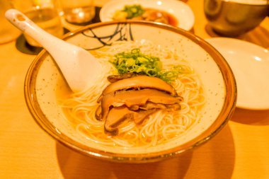 Buharda pişmiş Japon eriştesi çorbası dilimlenmiş mantarlar ve taze soğanlarla kaplı, ahşap bir masada sıcak ortam aydınlatması altında kırsal bir seramik kasede servis ediliyor..