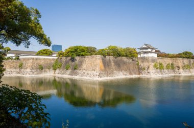 Büyük taş duvarlar ve Osaka 'daki açık mavi gökyüzünün altında yemyeşil yeşilliklerle çevrili sakin bir hendeğin üzerinde geleneksel bir Japon gözlem kulesi yükseliyor. Sakin su, tarihi mimariyi ve canlı ağaçları yansıtıyor. Huzurlu bir manzara yaratıyor..
