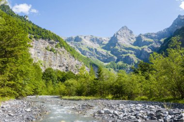 Kireçtaşı tepeleri ve Cheval Sirki 'ndeki engebeli zirvelerle çerçevelenmiş, kayalık bir yeşil ormanın içinden akan temiz bir dağ nehri. Parlak güneş ışığı ve mavi bir gökyüzü canlı yaz manzarasını güçlendirir..