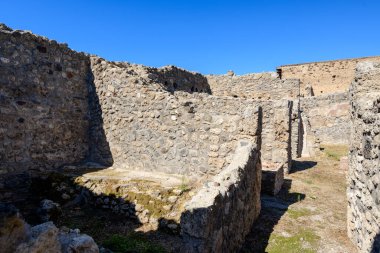 Pürüzlü volkanik taş duvarlar, İtalya 'nın Pompeii kentindeki eski yerleşim yerlerinde dar bir geçit ve açık bir alan oluşturur. Parlak güneş ışığı ve parlak mavi bir gökyüzü, arkeolojik alandaki yıpranmış duvarcılık ve dokulu zemini vurgular..