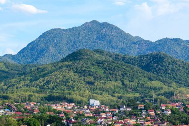 Luang Prabang, Laos 'un çatılarının arkasında yoğun yeşil tepeler yükseliyor, açık mavi gökyüzünün altında dramatik bir dağ zemini var. Sahne, canlı tropikal bitki örtüsü ile aşağıdaki kümelenmiş şehir arasındaki zıtlığı vurgular..