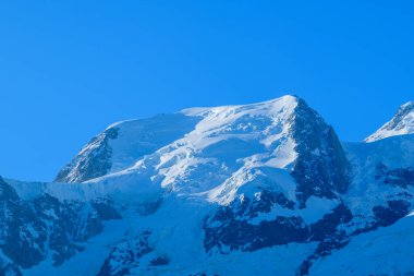 Mont Blanc du Tacul 'un karla kaplı zirvesi canlı mavi bir gökyüzüne karşı duruyor, pürüzsüz yamaçları ve engebeli kaya yüzeyleri Fransız Alpleri' nin sabah güneşiyle aydınlanıyor..