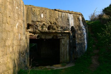 Yeşil çimenler ve açık mavi gökyüzünün altında kısmen açık bir girişi olan yıpranmış beton bir sığınağın yakın görüntüsü. Kaba, dokulu yüzey ve açısal yapı tarih ve terk edilme duygusunu çağrıştırıyor..