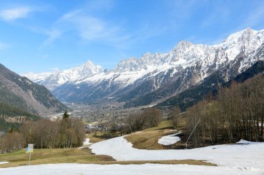 Dağınık bahar karları, çıplak ağaçlar ve parlak mavi gökyüzünün altındaki dramatik kar kaplı Mont Blanc yığını ile Chamonix vadisini süpüren manzara..