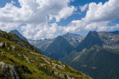 Sarp dağ zirveleri yemyeşil yamaçların ve kayalık çıkıntıların üzerinde yükseliyor, aşağıdaki orman parçalarıyla ve dalgalanan bulutlar canlı mavi gökyüzünde sürükleniyor..