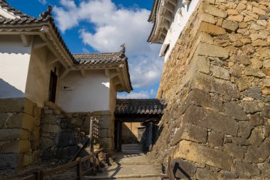 Himeji Şatosu 'nun tarihi bir taş duvar ve ahşap kapısının açılı görüntüsü, dokulu duvarlar, beyaz alçı duvarlar ve dramatik güneş ışığı mavi gökyüzünün altında güçlü gölgeler bırakıyor..