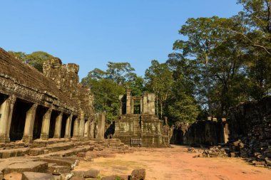 Altın öğleden sonra ışığı, Angkor, Kamboçya 'daki Bayon Tapınağı' nın açık avlusunu aydınlatıyor ve yıpranmış taş sütununu aydınlatıyor. Antik kumtaşı yapılar, yüksek orman ağaçlarının ve dağılmış duvarların arasında dururlar berrak mavi gökyüzünün altında..