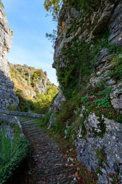 Yunanistan 'ın Epirus kenti Pont Gefiri Kokkorou yakınlarındaki Vikos Vadisi' ndeki dik kireçtaşı kayalıkları ve yemyeşil alanlar arasında uzanan eğimli taş patika. Yumuşak gün ışığı kanyon yolu boyunca dokulu kaya yüzeylerini ve doğal bitki örtüsünü vurgular.