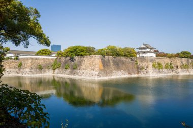 Geleneksel Japon kalesi tareti, Osaka 'da berrak mavi bir gökyüzü altında yemyeşil yemyeşil bir hendekle çevrili devasa taş surların ve sakin bir hendeğin üzerinde yükselir. Yansımalar sakin suda dans eder, barışçıl ve zamansız bir atmosfer yaratır..