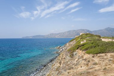 Cala Caterina 'daki berrak turkuaz suların üzerinde yeşil çalılarla kaplı güneşli kayalık kayalıklar yükseliyor. Uzak dağlar ve parlak mavi bir gökyüzü, huzurlu bir Akdeniz manzarası yaratıyor..