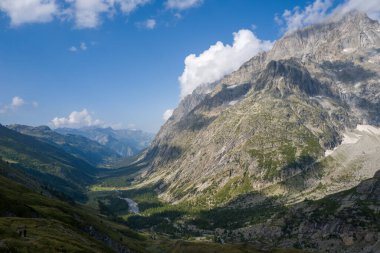 Val Ferret 'in dramatik manzarası. Yükselen kayalık dağlar, aşağıdaki dolambaçlı bir nehir ve yemyeşil yamaçların üzerine düşen gölgeler canlı mavi bir gökyüzü..