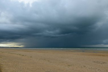 Ouistreham 'daki Kılıç Sahili' nin engin kumlu kıyı şeridi dramatik, karanlık bir yağmur fırtınasının altında ağır bulutların Manş Denizi 'ni kapladığı ve gergin ve dengesiz bir deniz manzarası oluşturduğu yerde..
