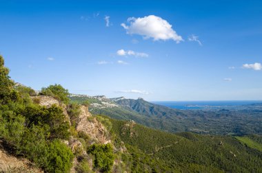 Sardunya 'nın Baunei ve Dorgali yakınlarındaki mavi Akdeniz manzaralı yuvarlanan tepelerin üzerinde sivri kayalar ve yoğun yeşil çalılar yükseliyor. Parlak güneş ışığı ve dağınık bulutlar canlı ve açık bir atmosfer yaratır..