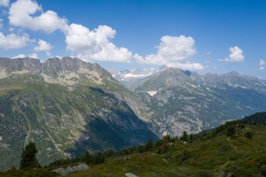 Yoğun ormanlarla çevrili sarp yeşil dağlar ve Emosson bölgesinde dağınık bulutlarla dolu canlı mavi bir gökyüzünün altındaki engebeli tepeler arasında uzanan beton bir barajın süpürme görüntüsü..