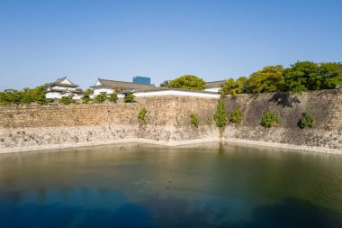 Geleneksel Japon köşe kuleleri ve yükseltilmiş taş duvarlar Osaka 'da sakin bir hendeğin üzerinde yükseliyor, manikürlü yeşillik ve açık mavi gökyüzü ile çevrili. Parlak güneş ışığı tarihi mimariyi ve sakin su yüzeyini vurguluyor.