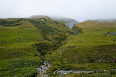 Yuvarlanan yeşil tepeler ve kayalık bir dere rüzgarı alçak sis ve bulutlardan oluşan bir battaniyenin altında sakin bir alp otlağının arasında. Çimen ve taşın ince dokuları barışçıl ve doğal bir atmosfer yaratır..