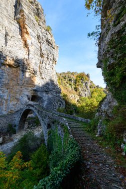 Historic arched stone bridge winds across a rocky gorge below towering limestone cliffs and lush autumn foliage near Pont Gefiri Kokkorou in Epirus, Greece. Crisp daylight highlights textured masonry, vibrant greenery, and dramatic canyon scenery.