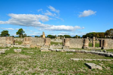 Tarihsel tuğla sütunlar ve yıpranmış taş duvarlar İtalya 'nın Paestum kentindeki arkeolojik sahanın çimenli açık zemininde duruyor. Sahne, dağınık bulutlar ve uzak ağaçlarla dolu canlı bir mavi Akdeniz gökyüzünün altındaki doğal güneş ışığıyla aydınlanıyor..