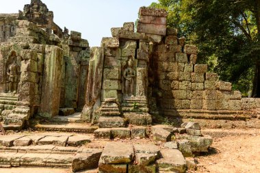 Antik yosun kaplı taş işçiliği ve karmaşık oymalar Kamboçya 'nın Angkor bölgesindeki Preah Khan tapınağının çevresini kaplıyor. Sahnede desenli bloklar, heykel şeklinde bir figür ve benekli güneş ışığı altında dağılmış kalıntılar yer alıyor.